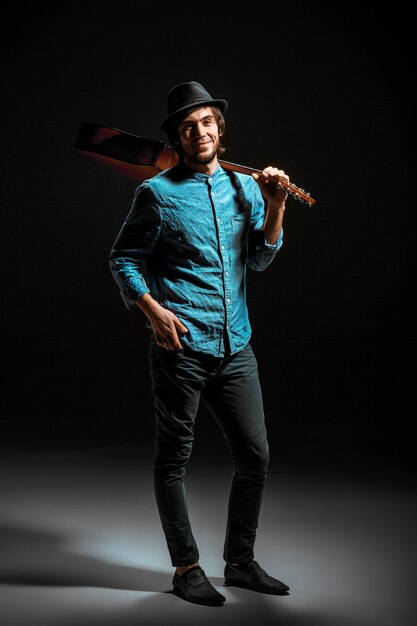 Cool guy with hat standing with guitar on dark studio background