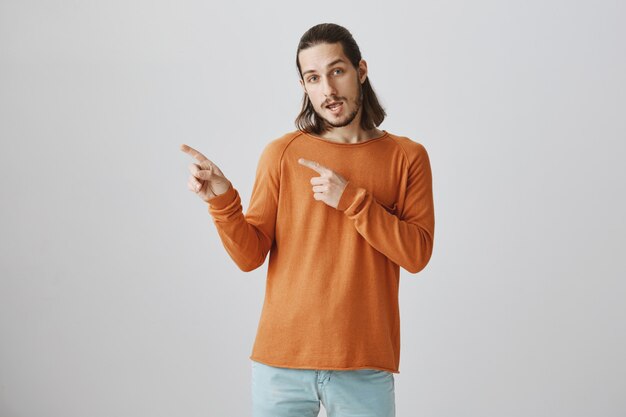 Cool handsome hipster guy pointing finger left, inviting to take look