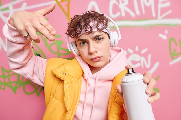 Cool handsome teenager dressed in hoodie and vest holds aerosol spray makes creative graffiti on street walls listens music via wireless headphones