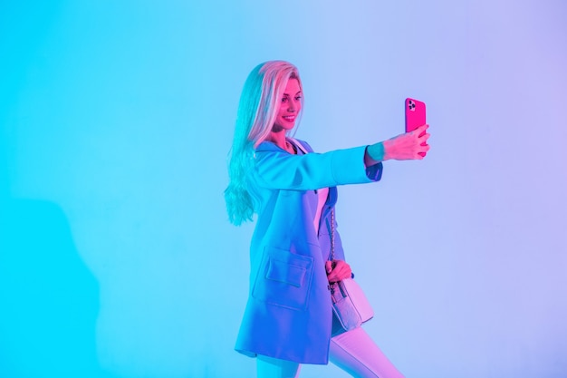 Крутая счастливая молодая деловая женщина в яркой модной одежде с пиджаком делает селфи на телефоне в студии на неоновом розовом светлом фоне
