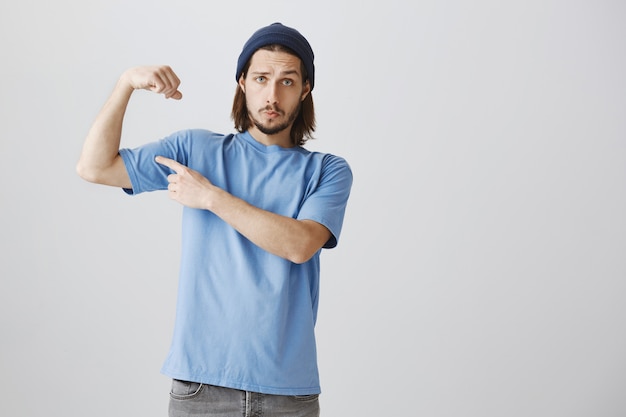 Крутой хипстерский парень в синей футболке и шапочке сгибает бицепсы, хвастаясь сильными мускулами
