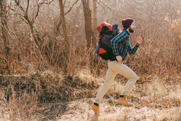 Cool hipster man traveling with backpack in autumn forest wearing checkered shirt and hat, active tourist running, exploring nature in cold season