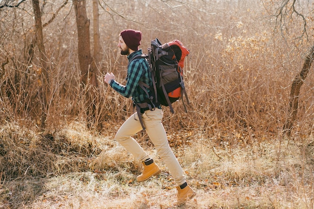Крутой хипстерский мужчина путешествует с рюкзаком в осеннем лесу в клетчатой рубашке и шляпе