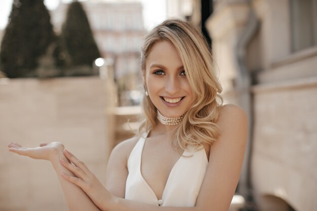 Крутая жизнерадостная блондинка в белом платье с v-образным вырезом и жемчужном ожерелье искренне улыбается, смотрит в камеру и указывает на улицу налево.