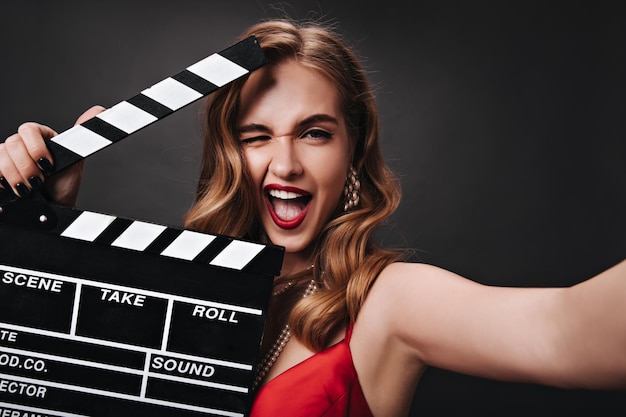 Cool lady in silk dress holding clapperboard and winking Emotional happy woman in red top laughing and posing in great mood on black backdrop