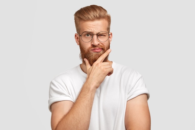 Cool man hipster with thick ginger hair, holds chin, blinks with eye, has fashionable hairdo