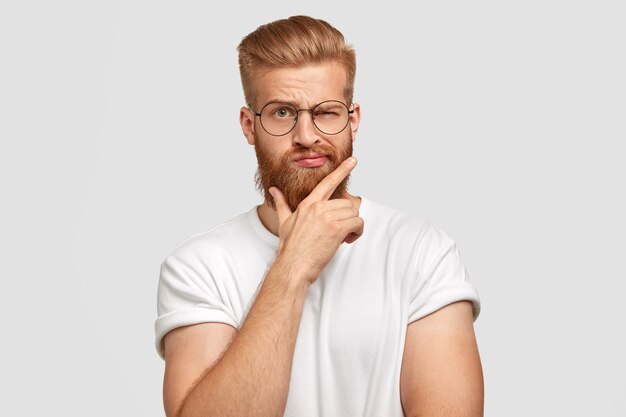 Cool man hipster with thick ginger hair, holds chin, blinks with eye, has fashionable hairdo