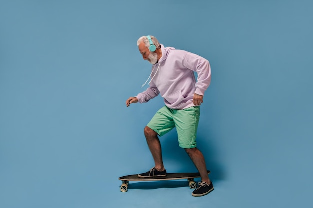 Cool man in hoodie listening to music in headphones Stylish adult guy with white beard in lilac clothes posing on isolated background