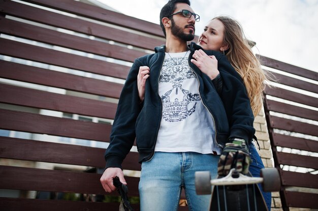 Cool multiracial couple hugs together with longboard against wooden fence