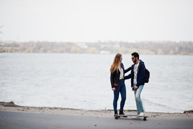 Cool multiracial couple standing with longboard at road against lake