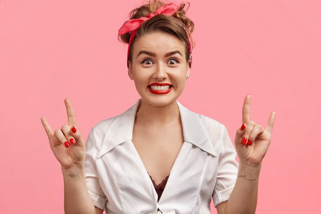 Cool pretty pinup woman with makeup, has happy expression, shows peace sign, gestures actively, wears retro clothing, has appealing appearance, isolated over pink