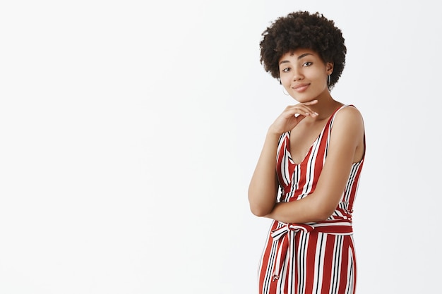 Cool self-assured and attractive dark-skinned african female coworker in stylish striped overalls, touching chin and smirking, crossing hand on chest, standing in confident pose over grey wall