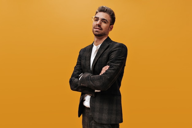 Cool self-confident man in checkered jacket and white t-shirt looks into camera. Bearded guy crosses arms and poses on orange wall.