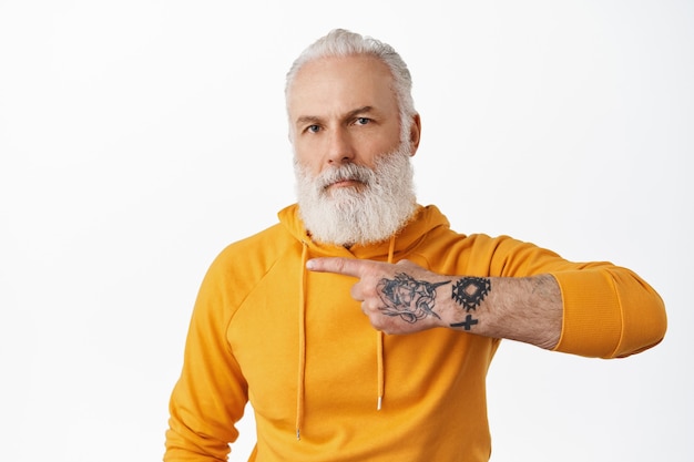 Cool senior hipster man with tattoos pointing left at copy space, showing advertisement, grandad with long beard wearing stylish hoodie recommending click link, white wall