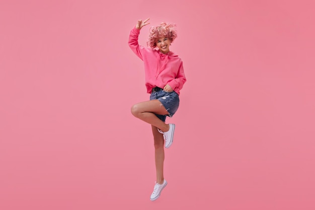 Cool shorthaired girl in pink hoodie and denim shorts jumping and smiling Happy woman with pink hair shows peace sign on isolated