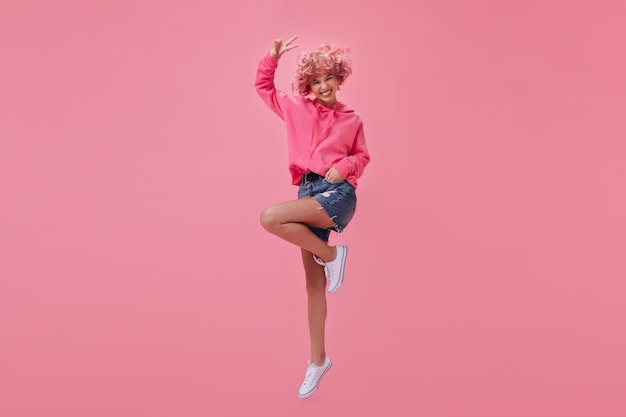 핑크색 까마귀와 데님 반바지를 입은 멋진 짧은 머리 소녀가 점프하고 웃고 핑크 머리를 가진 행복한 여자는 고립 된 상태에서 평화 기호를 보여줍니다