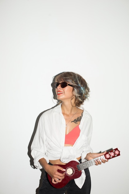 Cool smiling girl in white shirt and sunglasses happily looking aside while playing on little guitar over white background