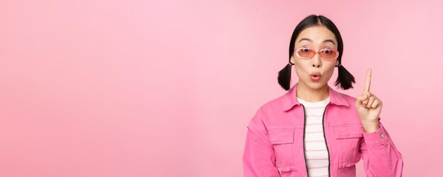 Cool and stylish asian girl raising finger pointing up suggesting smth pitching an idea or plan standing over pink background