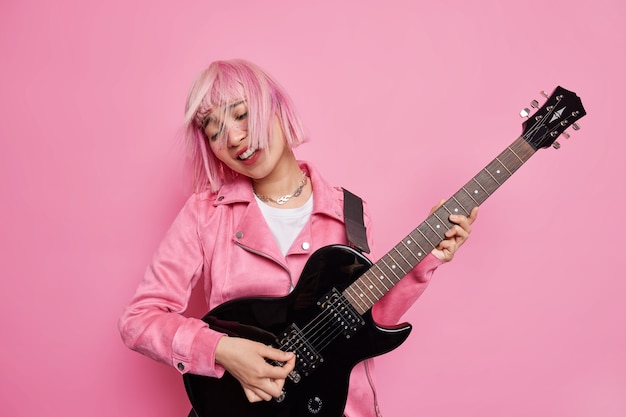 Free photo cool stylish female rocker plays famous song on electric guitar tilts head has pink hair floating on wind demonstrates her talent wears jacket