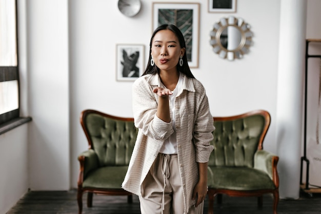 Cool tanned brunette lady in beige outfit blows kiss in cozy living room Happy Asian lady in cardigan and pants poses near beautiful green sofa