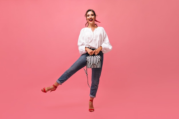 Cool woman in jeans and blouse holding bag on pink background.  Stylish girl with red lipstick and bandage on her hair is having fun..