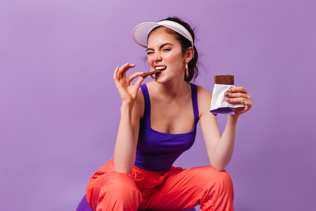 Foto gratuita cool woman in abbigliamento sportivo in stile anni '80 morde una deliziosa barretta di cioccolato al latte seduto sul muro viola