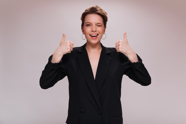 Cool woman winking and showing big thumbs up. Pretty girl in black jacket widely smiling and looking into camera on isolated background