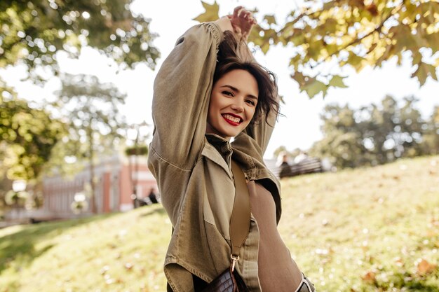 Cool woman with short hair in olive denim jacket smiling outside. Curly woman with bright red lipstick outdoors.