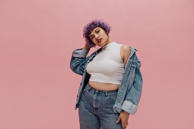 Cool woman with short multi-colored hair in round earrings, white T-shirt, large denim jacket and in light pants on pink.