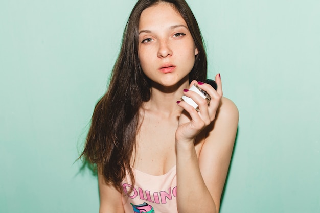 Cool young beautiful hipster woman posing against blue wall, holding bottle of parfume toilet water fragrance