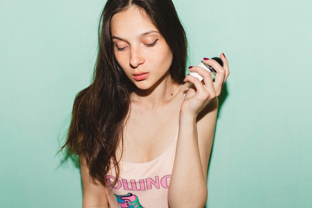 Cool young beautiful hipster woman posing against blue wall, holding bottle of parfume toilet water fragrance