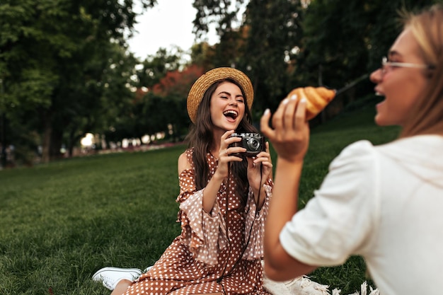 Cool young brunette woman in boater and polka dot dress laughs and takes photo of friend using retro camera Blonde girl in white outfit holds apple and has picnic Blurred effect