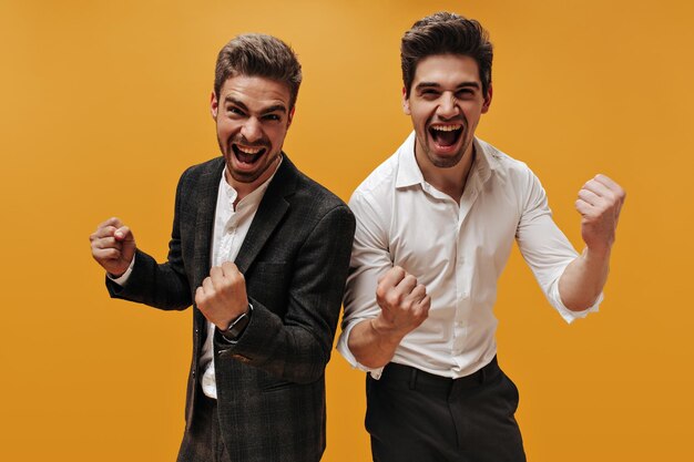 Cool young darkhaired businessmen in classical style outfits rejoice on orange background Excited guys in white shirts smile and scream on isolated
