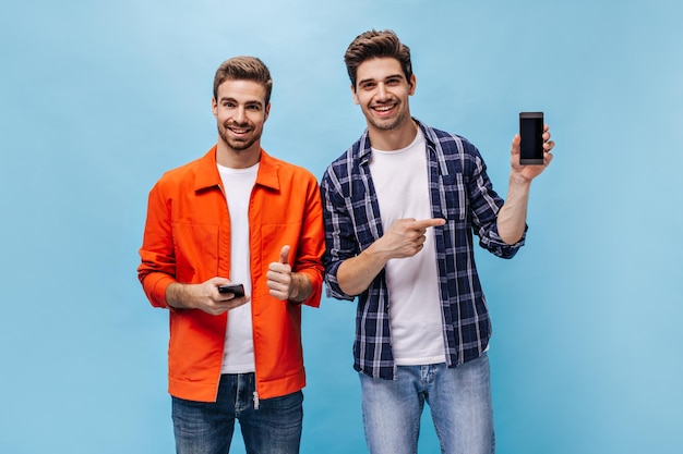 Free photo cool young excited men in stylish outfits pose on blue background guy in orange jacket shows thumb up man in checkered shirt points at phone screen