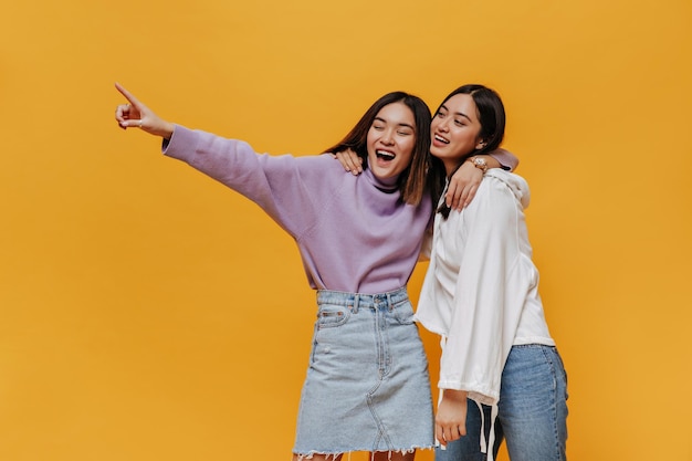 Cool young girl in purple sweater points at place for text on isolated Charming Asian women in good mood hug on orange background