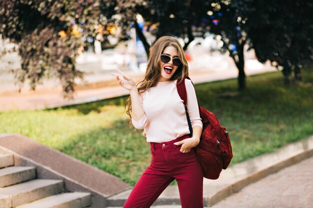 Cool young girl with vinous bag and long curly hair having fun in park in town. She wears marsala color and looks excited.