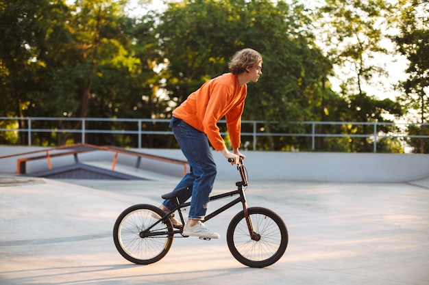 Cool young guy in orange pullover and jeans riding bicycle at modern skatepark