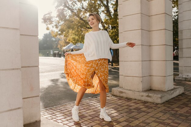 Cool young happy smiling woman in yellow printed dress and knitted white sweater on sunny autumn day having fun in street