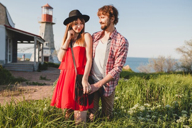 Cool young stylish couple in love in countryside, indie hipster bohemian style, weekend vacation, summer outfit, red dress, green grass, holding hands, smiling