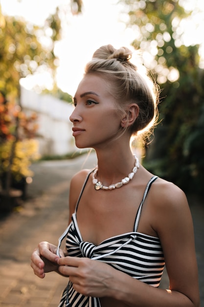 Free photo cool young tanned blonde woman with hair bun dressed in striped dress look away and walk in park