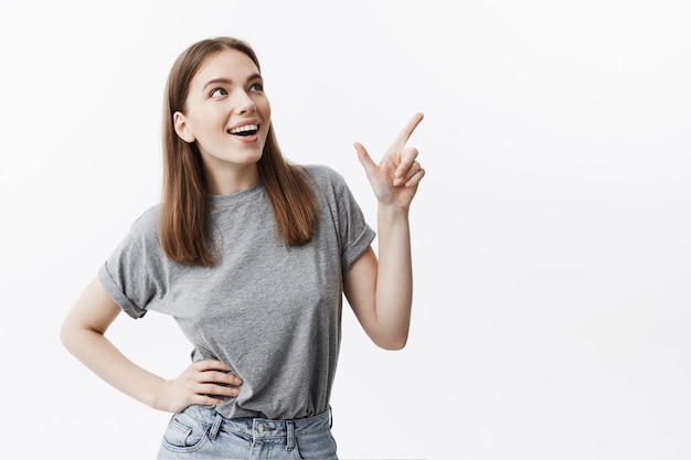 Copy space for advertisement. Close up of handsome caucasian young female student with dark hair in casual grey clothes smiling brightfully, looking aside with happy expression, pointing on white back