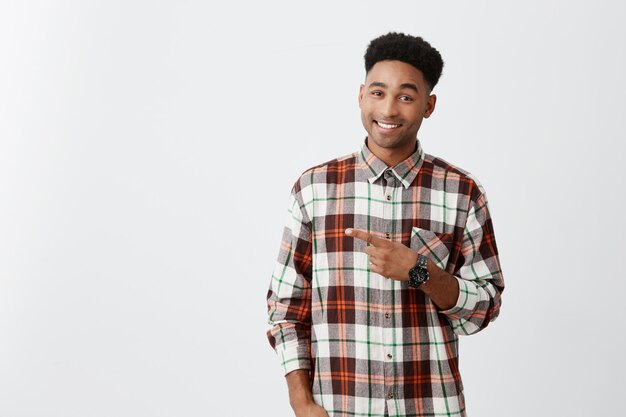 Copy space for advertisement. Dark-skinned young good-looking joyful man with curly hairstyle in checkered shirt pointing aside with one hand with smile and happy expression.