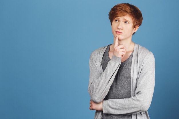 Copy space. Colorful blue wall Portrait of young beautiful male student in casual outfit holding finger near mouth with thoughtful expression, thinking about skipping lecture in university.