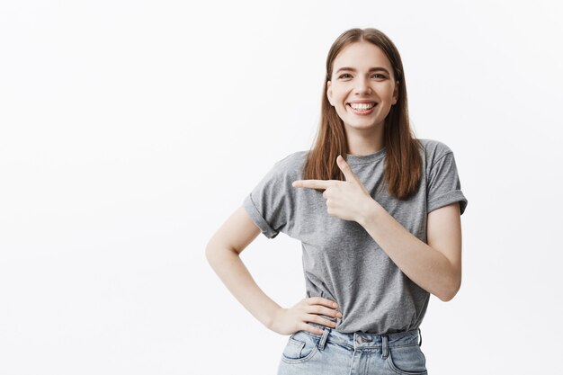 Copy space for your advertisement. Young beautiful caucasian dark-haired girl smiling,  with happy expression, pointing a side with finger on white wall.