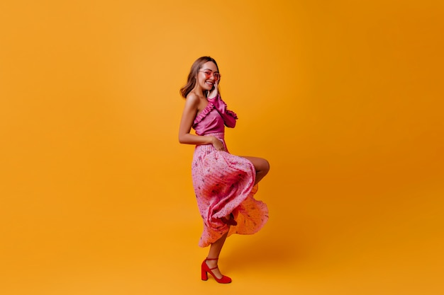 Coquette in lovely tender pink dress and red heels with joy on her face, posing in profile in warm