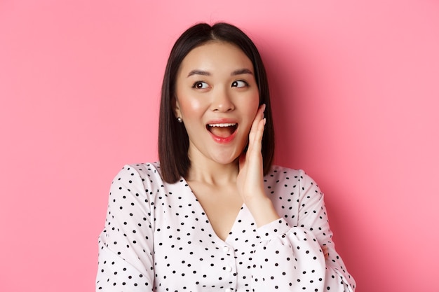 Coquettish asian woman in blushing touching cheek and looking left amused standing over pink background