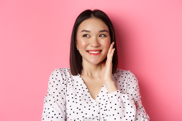Coquettish asian woman in blushing touching cheek and looking left amused standing over pink background