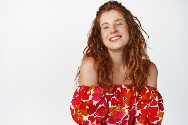 Coquettish redhead girl biting lip and looking flirty at camera posing in stylish floral dress standing against white background