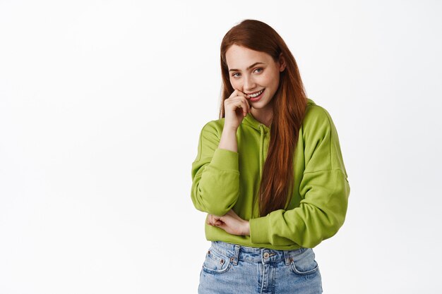 Coquettish and thoughtful redhead woman, smiling pleased and thinking, having coy, interesting idea, standing on white