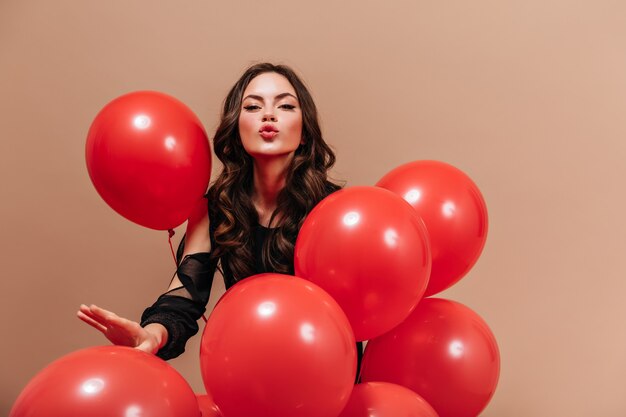 Coquettish woman in black blouse holds balloons and blows kiss on beige background.